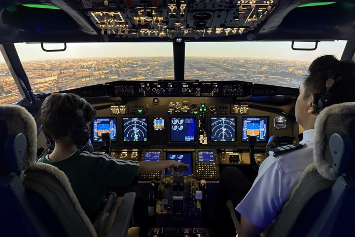 Child and pilot in airplane cockpit with city view outside.
