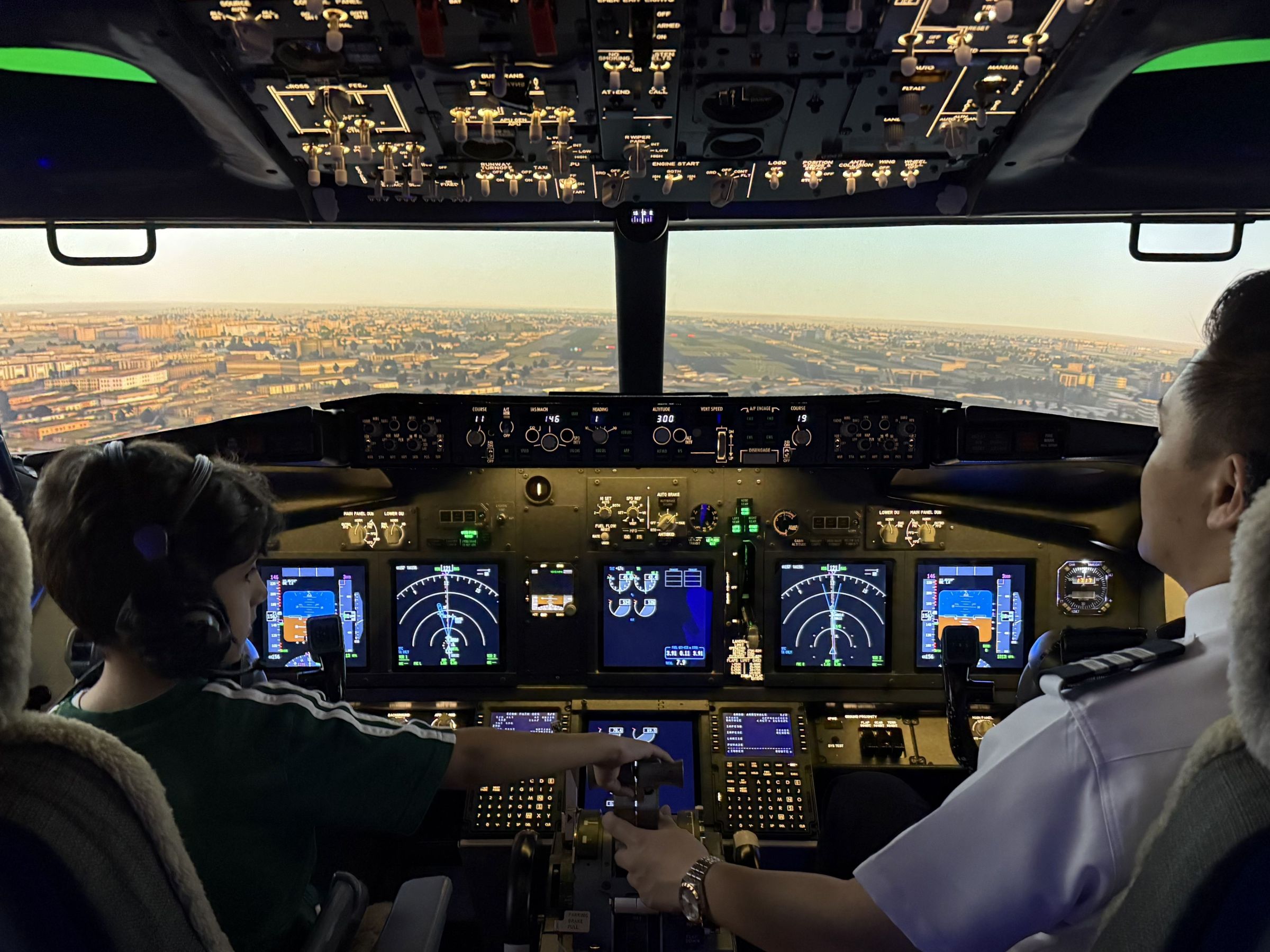 Two people operate an aircraft cockpit simulator with cityscape on display screen.